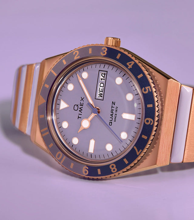 Q Timex® watch in rose gold-tone with a lilac dial and blue bezel, featuring a two-tone stainless steel bracelet with white center links, displayed against a soft lavender background.