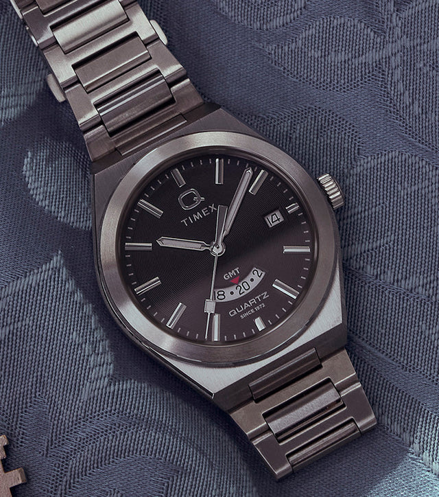 Q Timex® Continental GMT watch with a black dial, stainless-steel case, and bracelet displayed on a patterned blue fabric beside a vintage key and pink ticket stub.