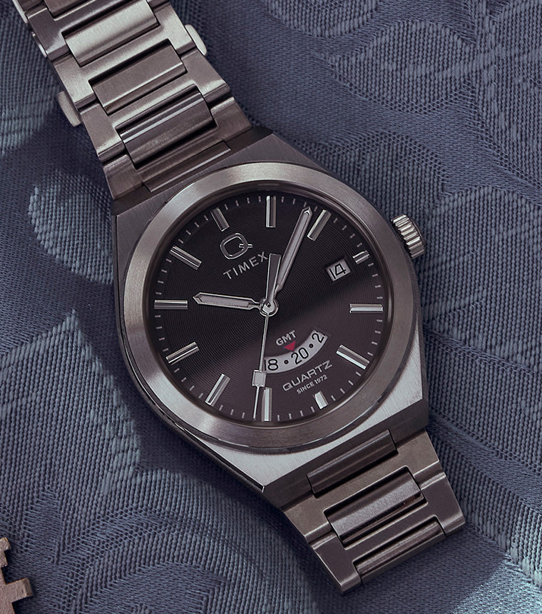 Q Timex® Continental GMT watch with a black dial, stainless-steel case, and bracelet displayed on a patterned blue fabric beside a vintage key and pink ticket stub.