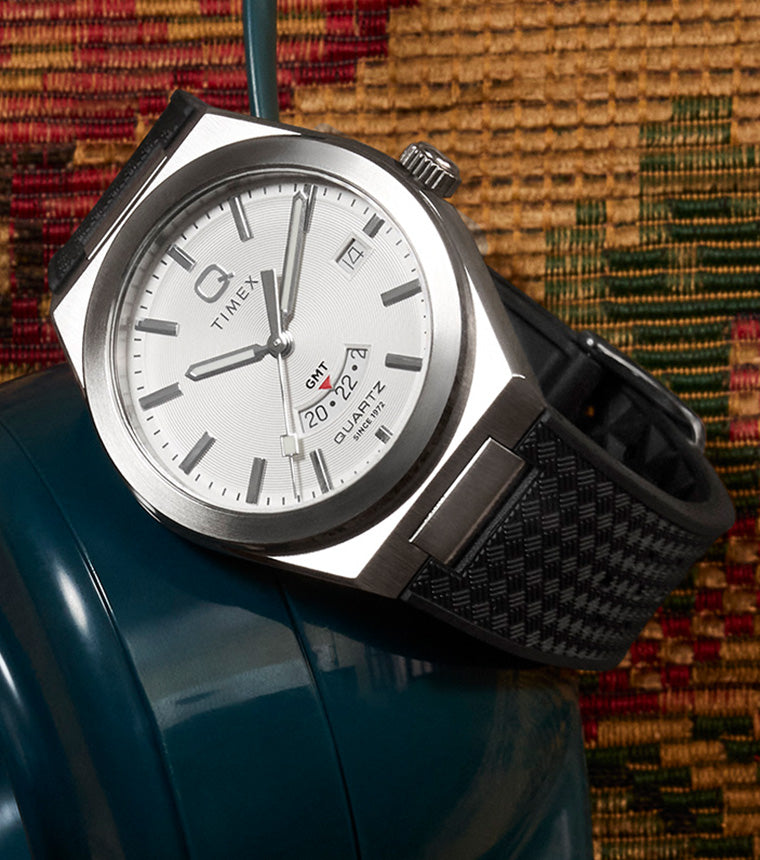 Q Timex® Continental GMT watch with a silver dial, stainless-steel case, and black woven strap displayed on a vintage tapestry background beside a teal rotary phone.