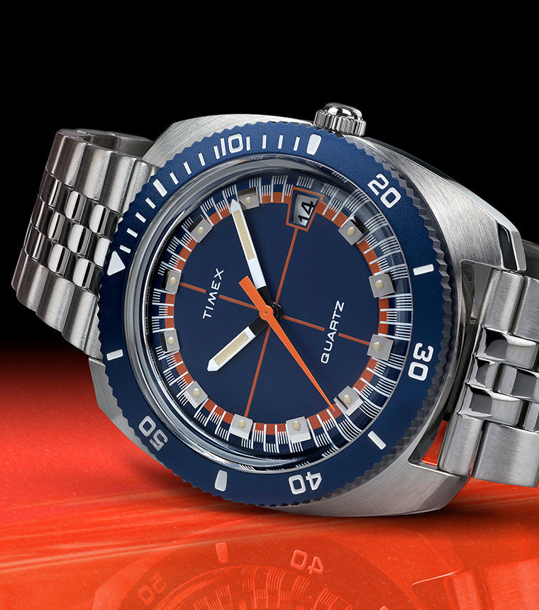 Q Timex® 1971 Velocity watch with a blue dial, orange accents, and stainless steel bracelet, displayed on a glossy red surface with a black background.