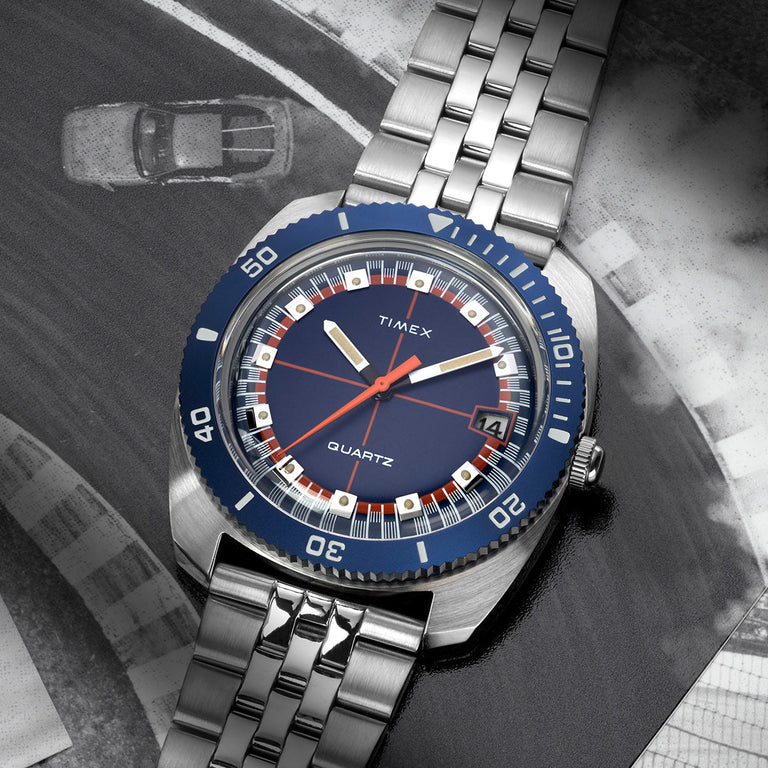 Q Timex® 1971 Velocity watch with a blue dial and silver bracelet, placed over a black-and-white racing track photo, emphasizing its vintage motorsport inspiration.