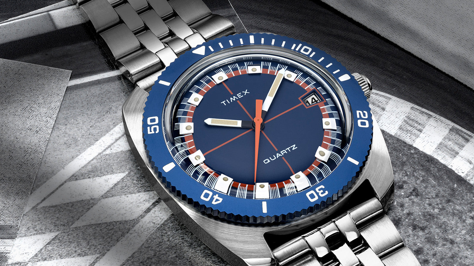 Detailed macro of the Q Timex® 1971 Velocity watch face, featuring a navy blue dial, orange crosshair design, and silver hour markers under a domed crystal.