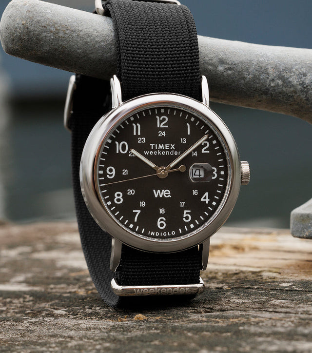 Timex Weekender watch with black dial, silver-tone case, and black fabric strap resting on a weathered wooden dock beside a metal cleat.