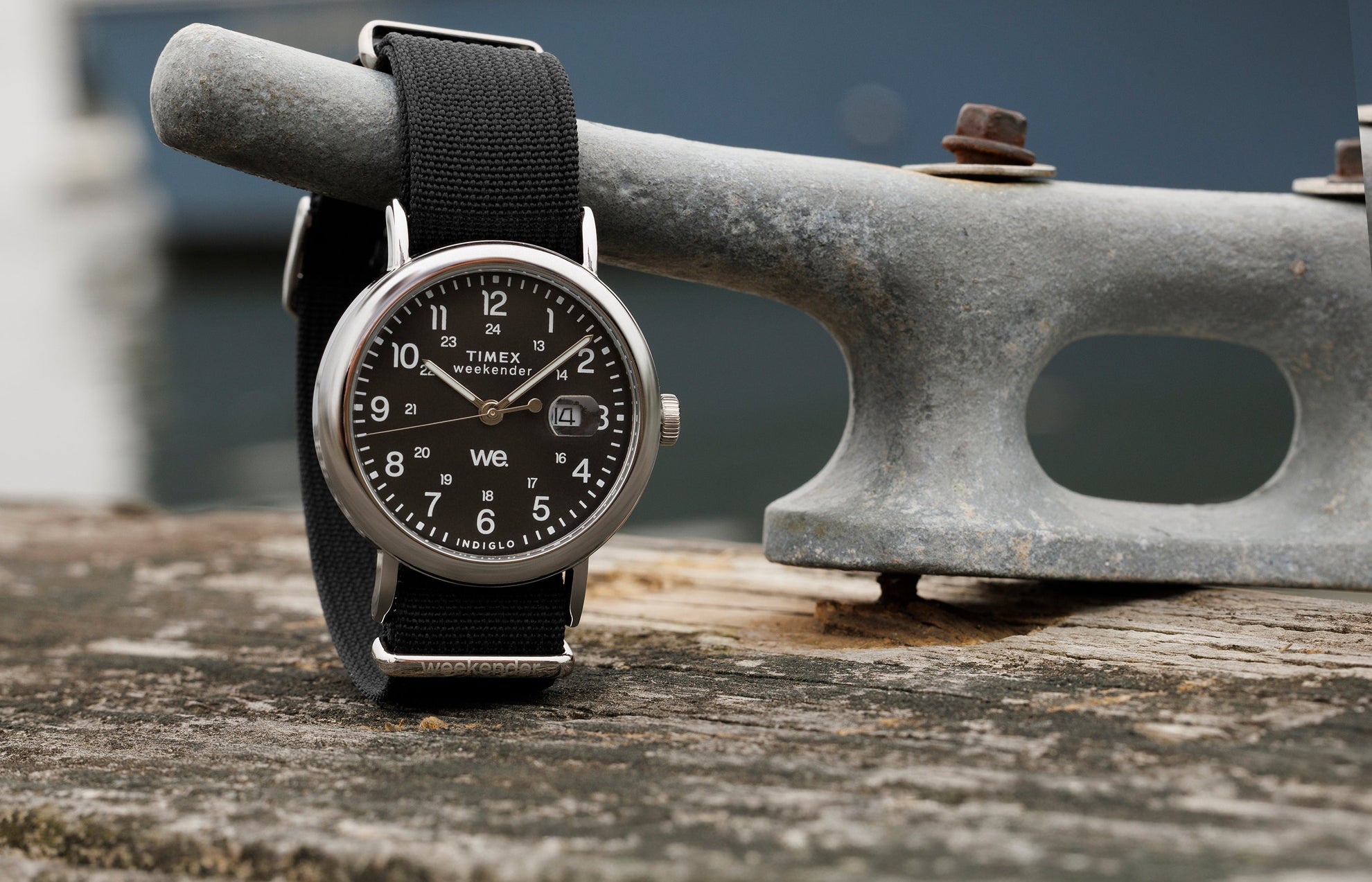 Timex Weekender watch with black dial, silver-tone case, and black fabric strap resting on a weathered wooden dock beside a metal cleat.