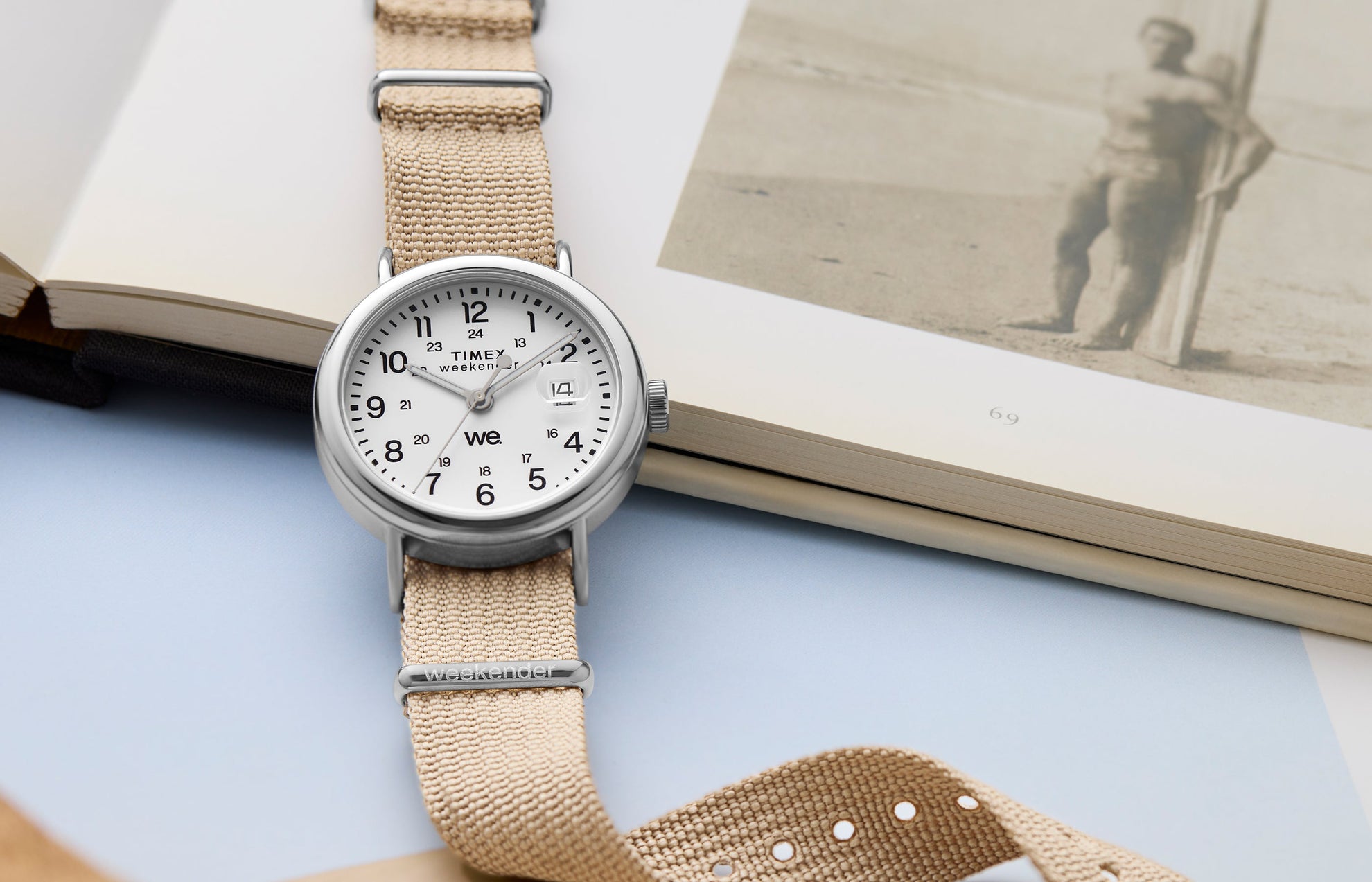 Timex Weekender watch with a beige fabric strap and white dial, placed beside an open book showing a vintage beach photograph.