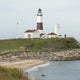 A coastal lighthouse with red-roofed buildings, grassy hill, and rocky shoreline along the ocean.