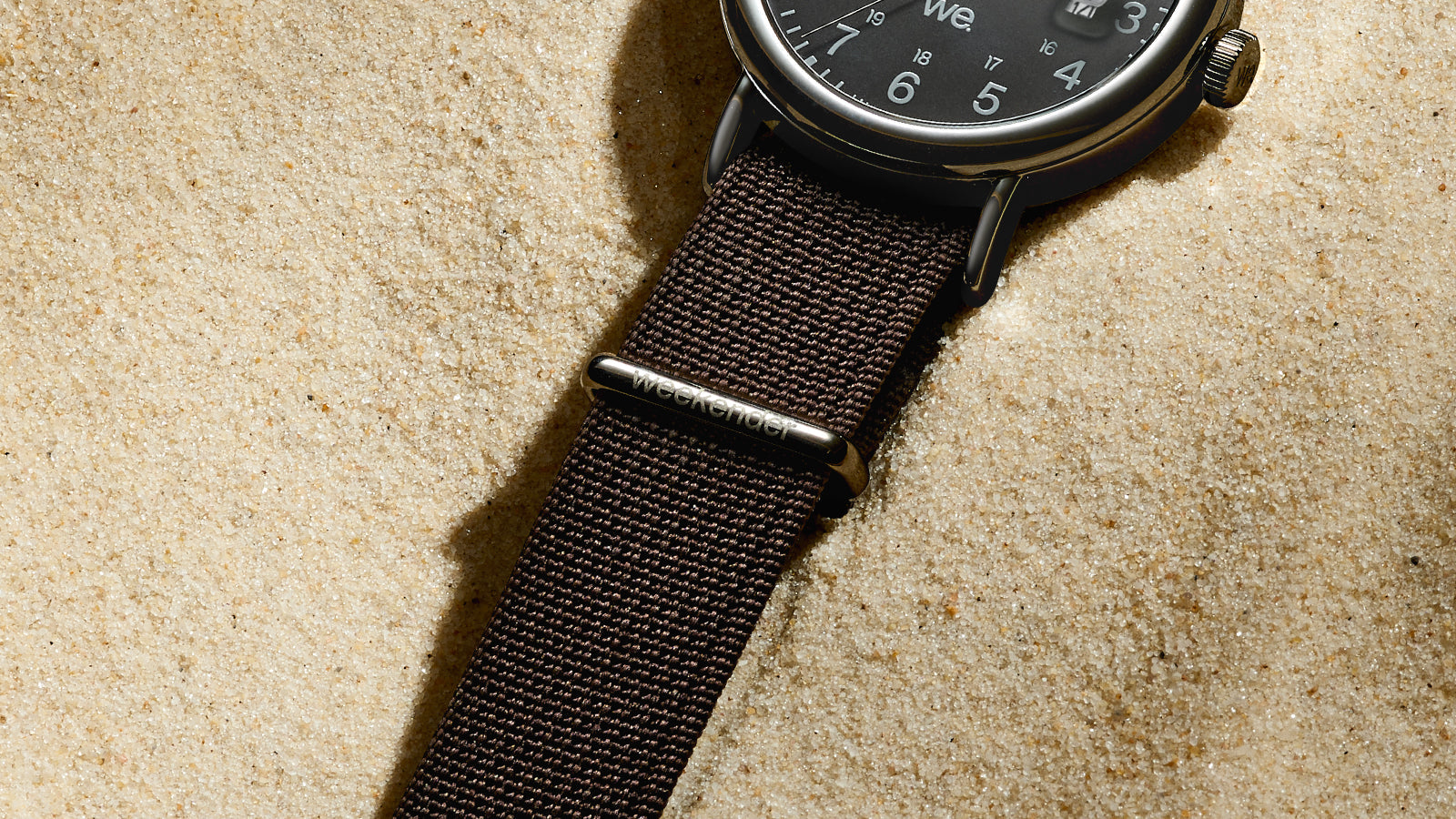 Close-up of the textured black fabric strap of the Timex Weekender with polished “weekender” branding on the loop, resting on sand.