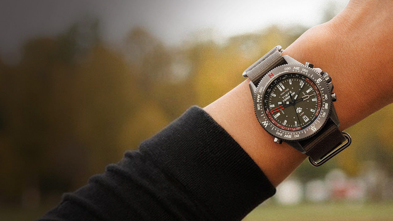 Timex Expedition Tide-Temp-Compass worn on the wrist, featuring a black dial, compass bezel, and green fabric strap, shown against a soft outdoor background.