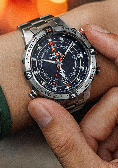 Close-up of a Timex Expedition Tide-Temp-Compass watch on a wrist, with a stainless-steel bracelet and multifunction black dial featuring tide, temperature, and compass indicators. The glowing firelight in the background highlights the rugged, outdoor-ready design.