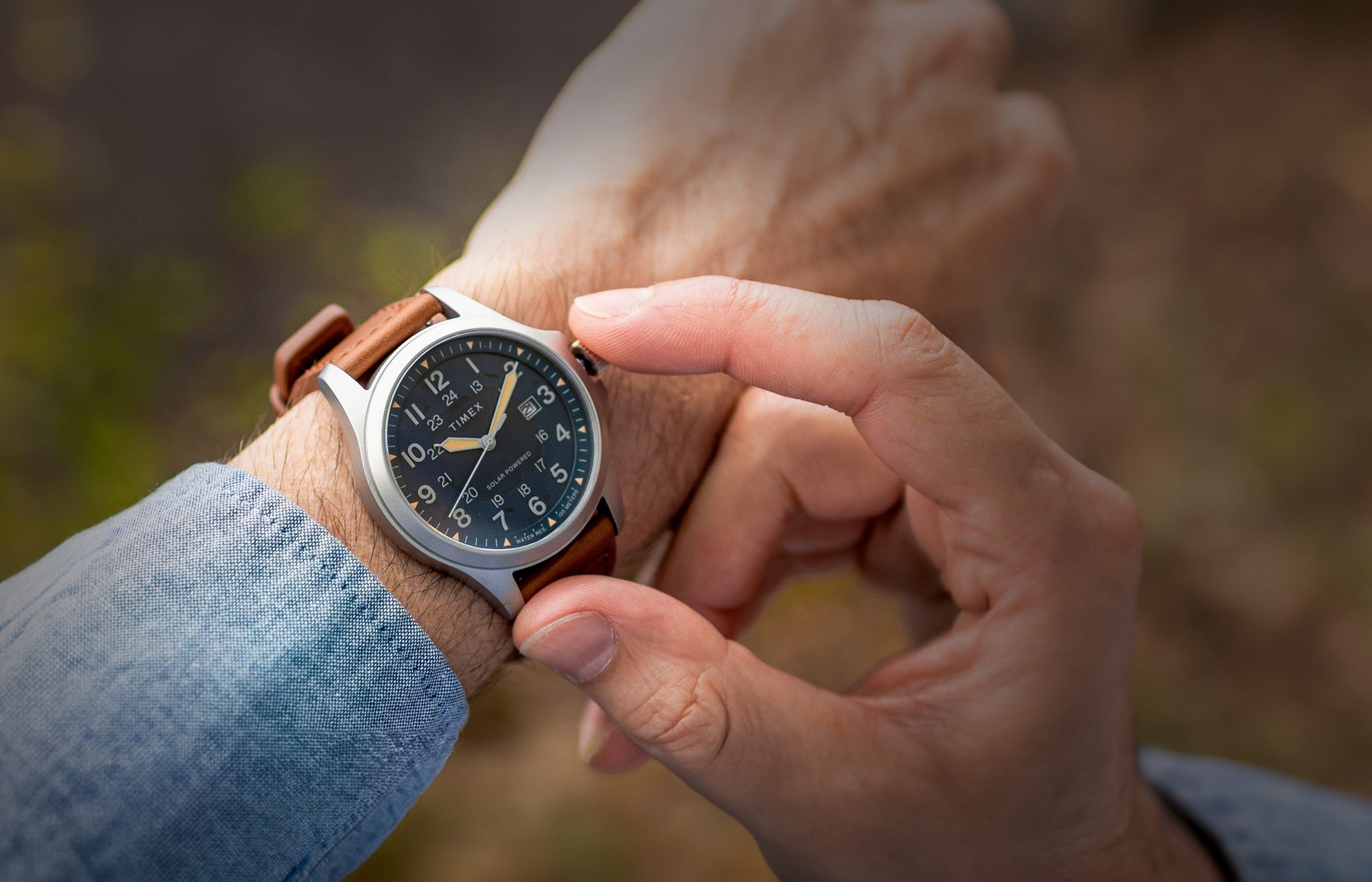 A Timex Expedition Field Post Solar watch with a navy dial and brown leather strap worn on the wrist, while the wearer adjusts the crown outdoors.