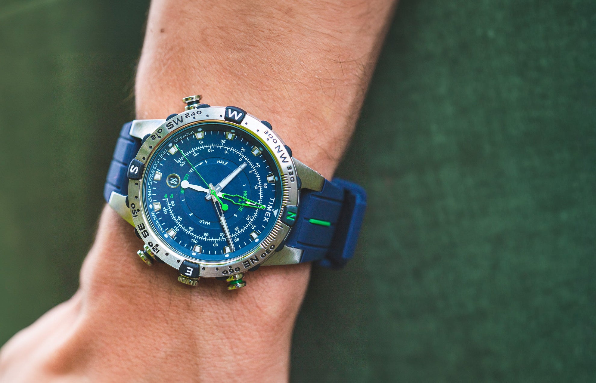 A Timex Expedition Tide-Temp-Compass watch with a blue dial and strap, worn on a wrist outdoors. The stainless-steel case features a rotating compass bezel, luminous hands, and green tide and temperature indicators.