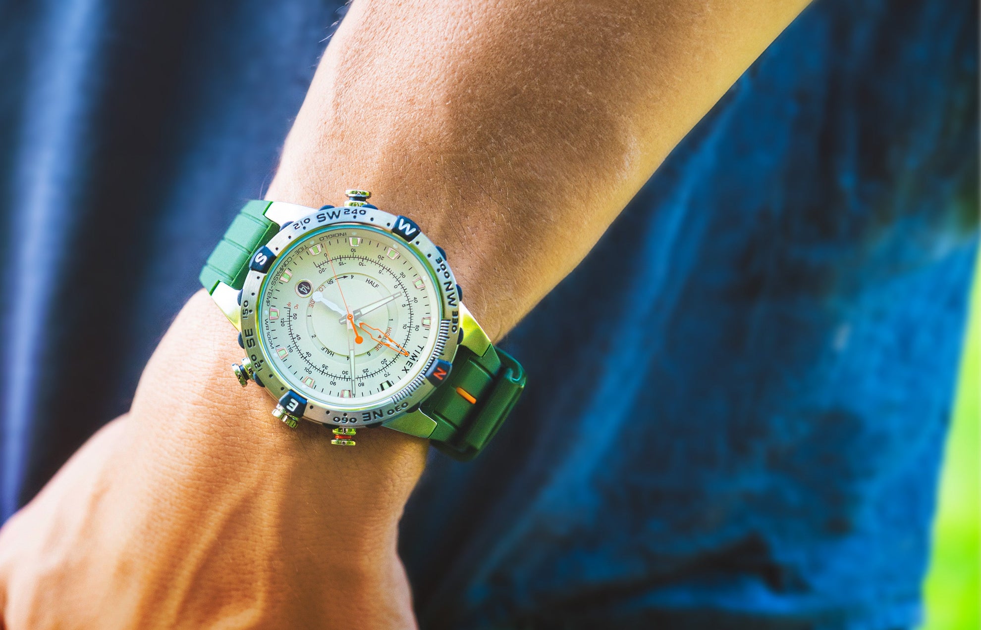 A Timex Expedition Tide-Temp-Compass watch with a cream dial, stainless-steel case, and green silicone strap worn on the wrist outdoors. Features luminous hands, orange tide indicators, and compass bezel accents.