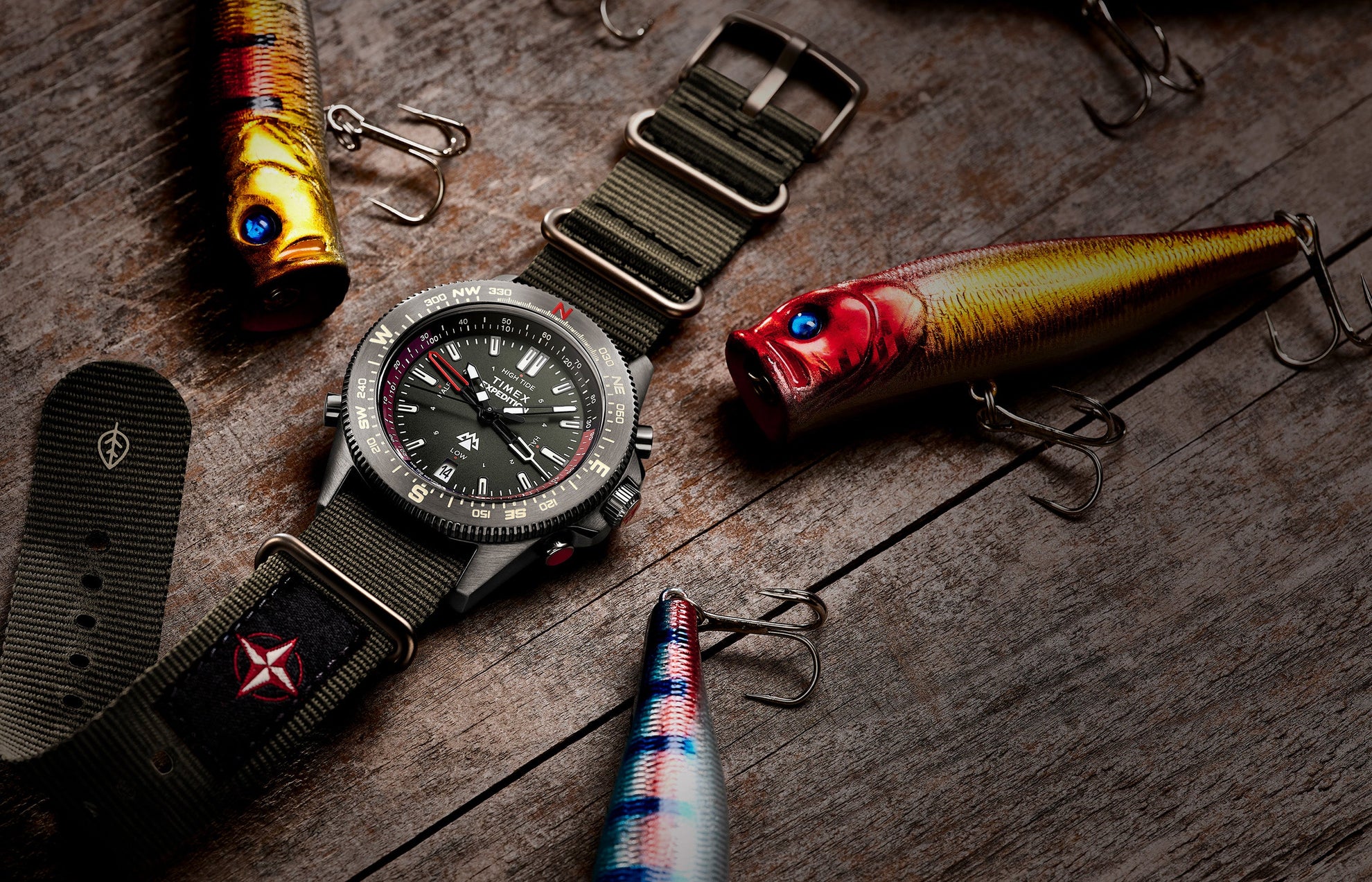 Timex Expedition Tide-Temp-Compass with green fabric strap displayed on a wooden surface among colorful fishing lures, highlighting its rugged outdoor style.