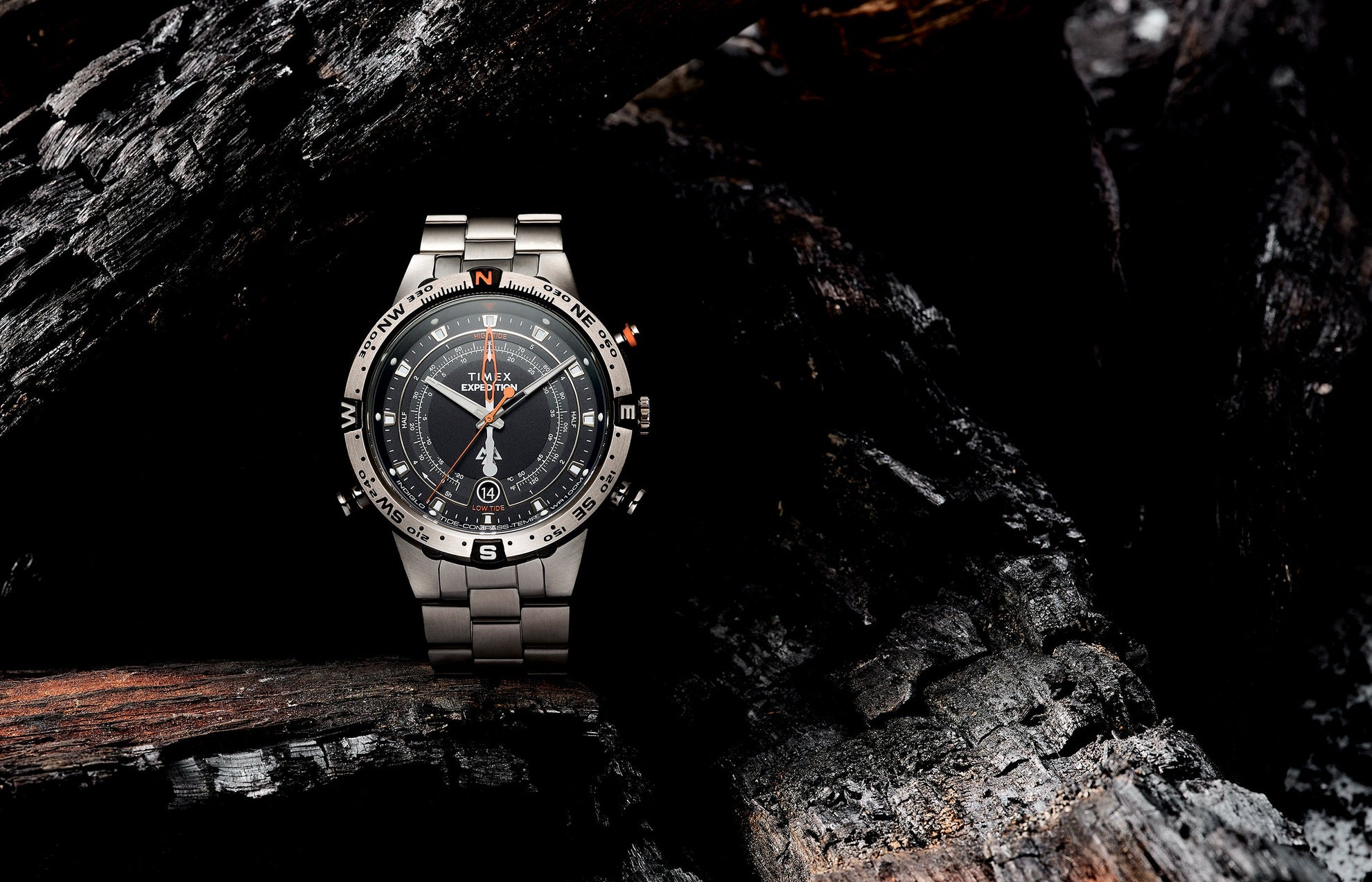 Timex Expedition Tide-Temp-Compass watch displayed on charred wood, showcasing its brushed stainless-steel bracelet, rotating compass bezel, and detailed black dial with orange accents.