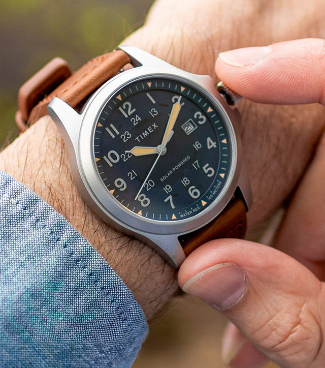 A Timex Expedition Field Post Solar watch with a navy dial and brown leather strap worn on the wrist, while the wearer adjusts the crown outdoors.