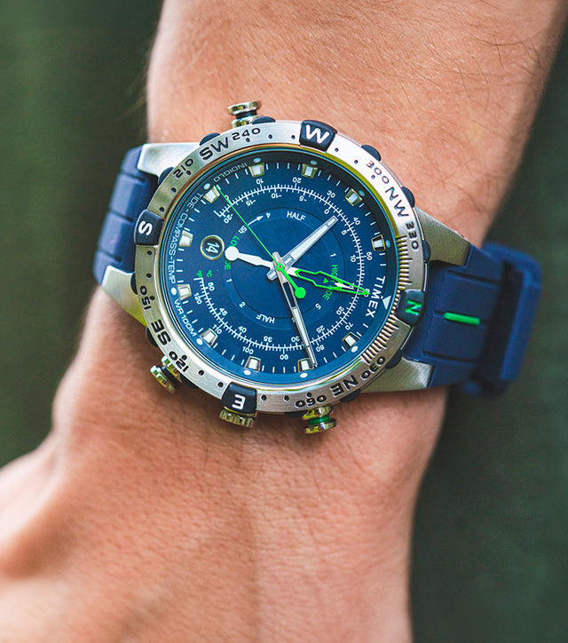A Timex Expedition Tide-Temp-Compass watch with a blue dial and strap, worn on a wrist outdoors. The stainless-steel case features a rotating compass bezel, luminous hands, and green tide and temperature indicators.