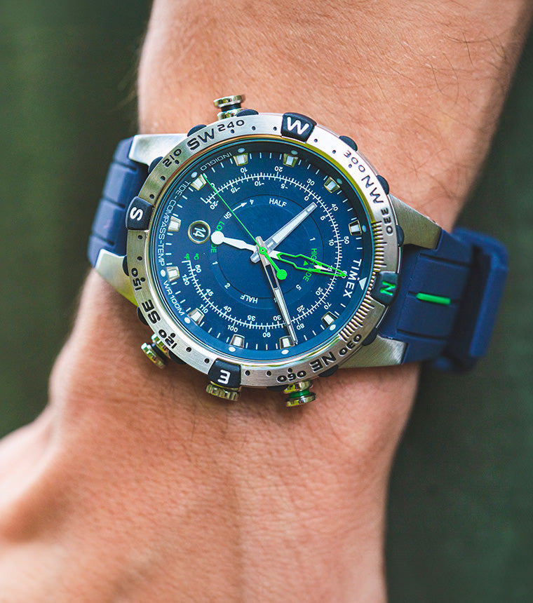 A Timex Expedition Tide-Temp-Compass watch with a blue dial and strap, worn on a wrist outdoors. The stainless-steel case features a rotating compass bezel, luminous hands, and green tide and temperature indicators.