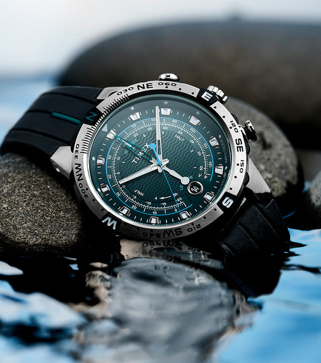 Close-up of the Timex Expedition Tide-Temp-Compass with a teal wave-pattern dial resting on smooth rocks beside water, showcasing its rugged, water-resistant design.