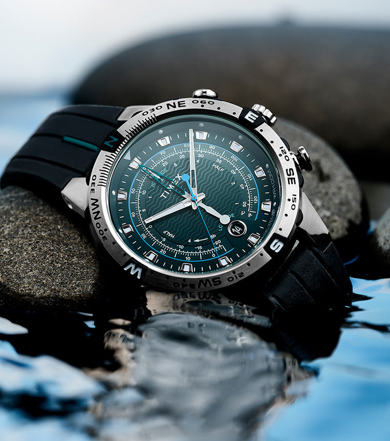 Close-up of the Timex Expedition Tide-Temp-Compass with a teal wave-pattern dial resting on smooth rocks beside water, showcasing its rugged, water-resistant design.