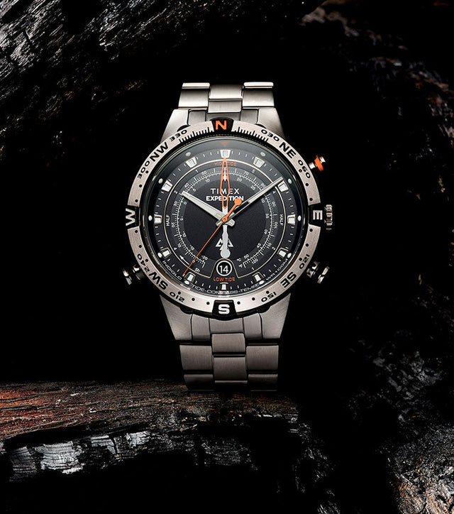 Timex Expedition Tide-Temp-Compass watch displayed on charred wood, showcasing its brushed stainless-steel bracelet, rotating compass bezel, and detailed black dial with orange accents.