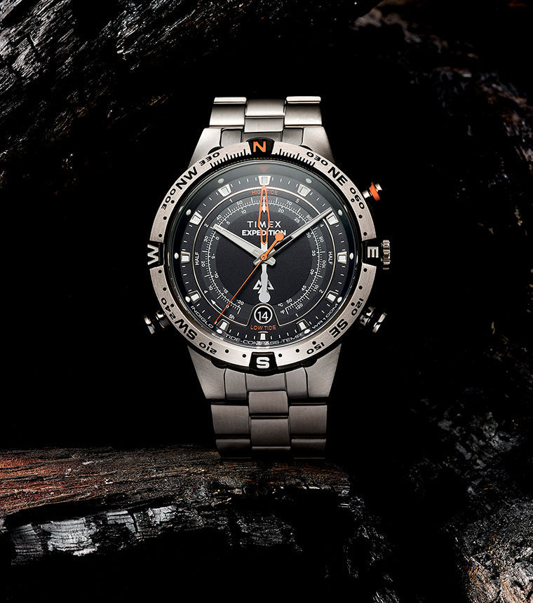 Timex Expedition Tide-Temp-Compass watch displayed on charred wood, showcasing its brushed stainless-steel bracelet, rotating compass bezel, and detailed black dial with orange accents.