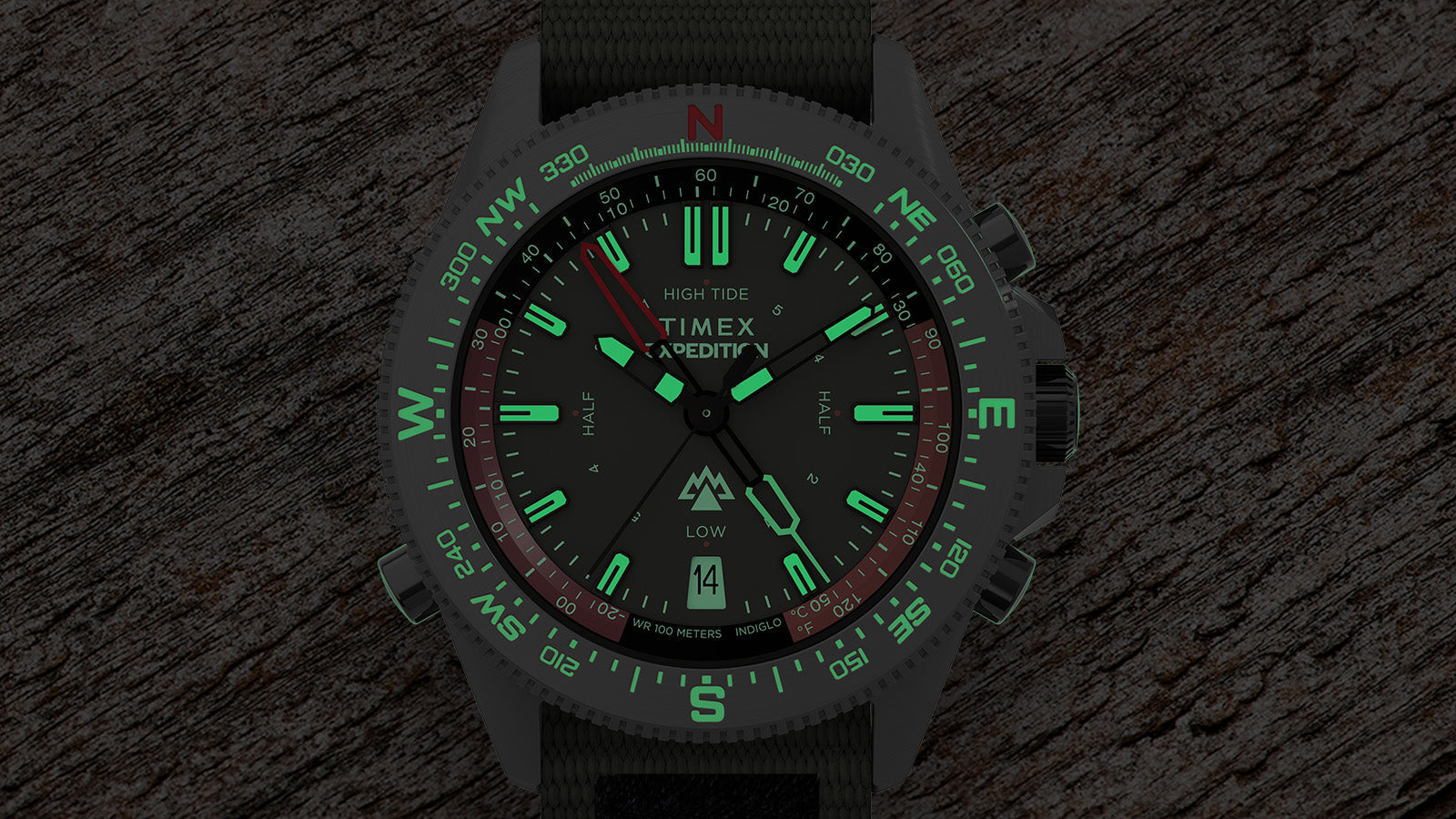 Timex Expedition Tide-Temp-Compass glowing in the dark with luminous green markers and hands, compass bezel visible, laid against a wooden surface.