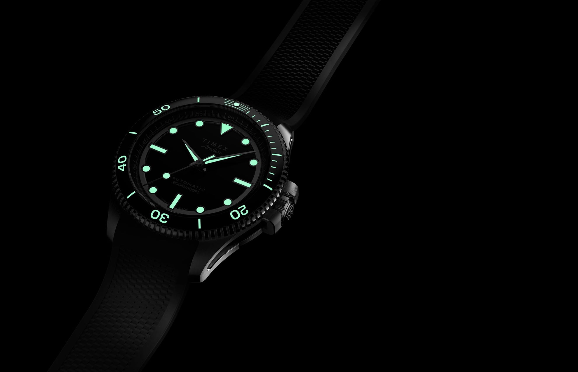 Timex Atelier dive watch glowing with bright green luminescent hands, indices, and bezel markers against a black dial, shown in the dark with rubber strap.