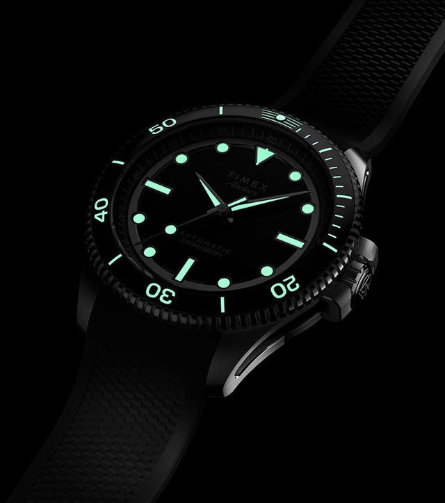 Timex Atelier dive watch glowing with bright green luminescent hands, indices, and bezel markers against a black dial, shown in the dark with rubber strap.