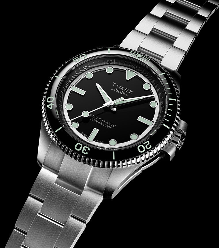 Timex Atelier automatic dive watch in brushed stainless steel with black dial, luminous markers, and unidirectional bezel, angled against a dark background.