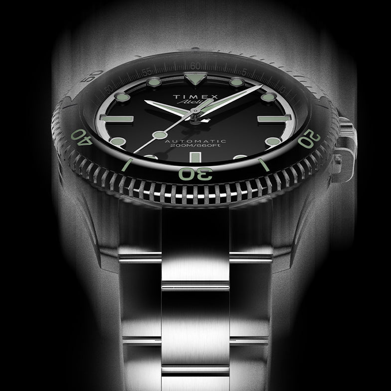 Front view of Timex Atelier automatic dive watch showing black dial, bold luminous indices, stainless steel bracelet, and engraved bezel detail.