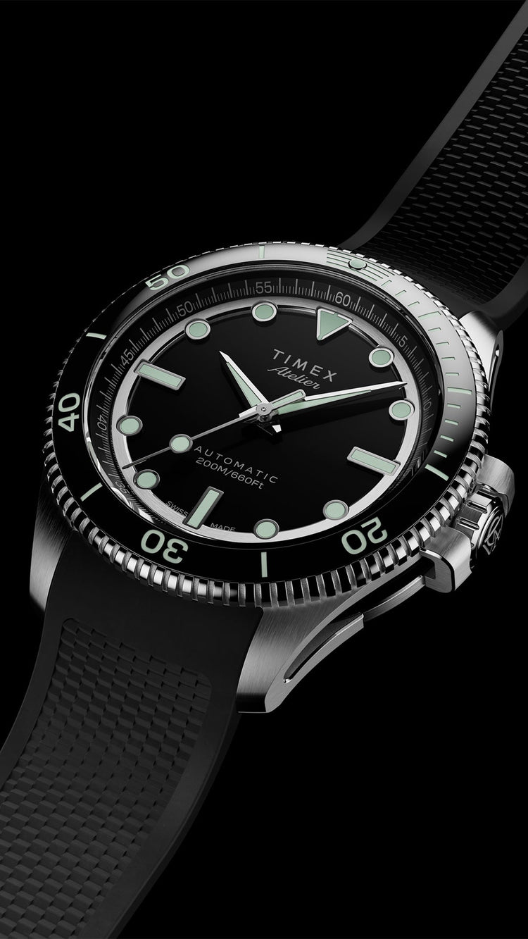 A Timex Atelier automatic dive watch with a black dial, luminous markers, rotating bezel, and a textured black rubber strap, shown in a close-up angled view against a black background.