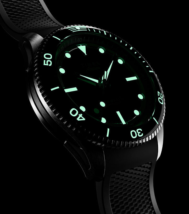 Timex Atelier automatic dive watch with black rubber strap shown in low light, highlighting the luminous green markers, bezel scale, and hands glowing brightly in the dark.