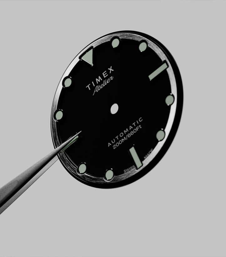 Close-up of the Timex Atelier watch dial being held with precision tweezers, displaying luminous indices, branding, and automatic 200M/660FT marking before assembly.