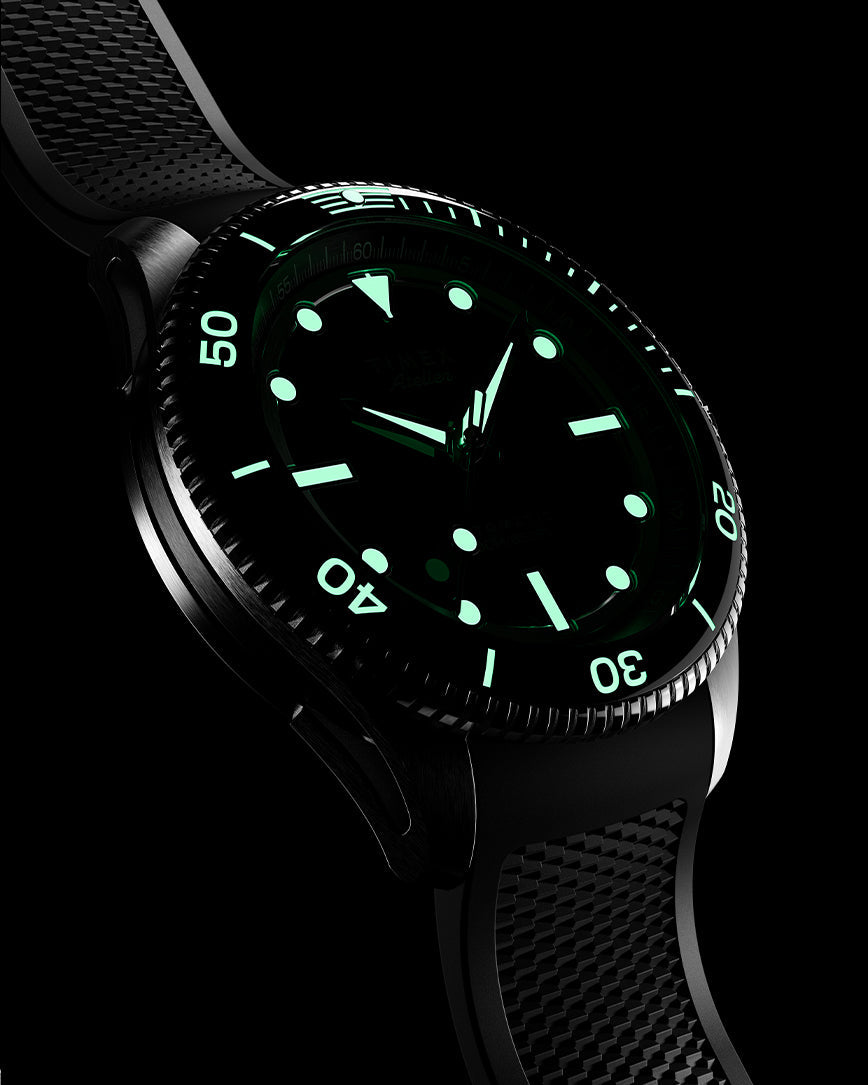 Timex Atelier automatic dive watch with black rubber strap shown in low light, highlighting the luminous green markers, bezel scale, and hands glowing brightly in the dark.