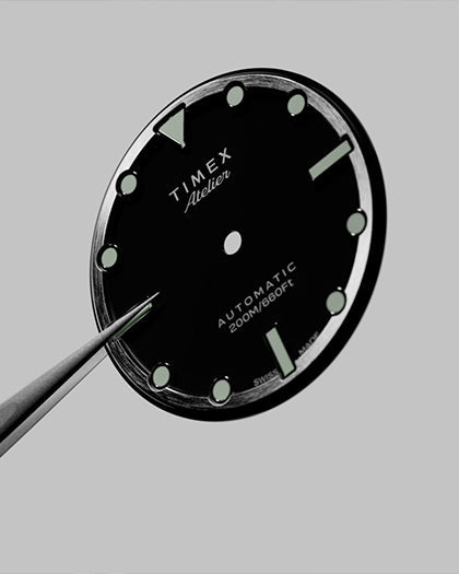 Close-up of the Timex Atelier watch dial being held with precision tweezers, displaying luminous indices, branding, and automatic 200M/660FT marking before assembly.