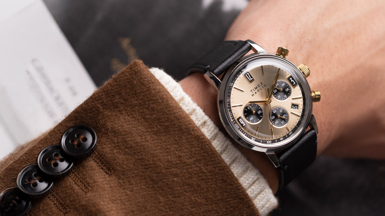 Timex Marlin Chronograph Tachymeter with champagne dial and black leather strap worn on wrist, paired with brown blazer and cream sweater cuff.