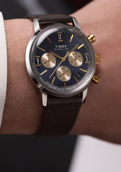 Timex Marlin Chronograph Tachymeter with navy blue dial, gold subdials, and brown leather strap displayed on blue background.
