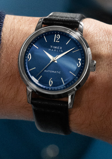 Timex Marlin Automatic watch with blue dial and silver-tone markers worn on wrist, paired with a brown jacket and navy knit cuff.