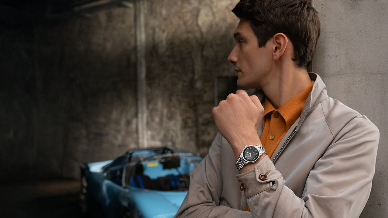 Model wearing Timex Marlin Automatic with black dial and stainless-steel bracelet, styled in beige jacket and orange shirt, standing beside a blue vintage car.