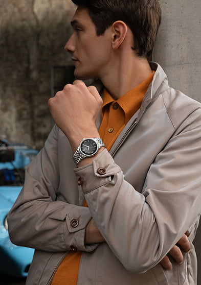 Model wearing Timex Marlin Automatic with black dial and stainless-steel bracelet, styled in beige jacket and orange shirt, standing beside a blue vintage car.