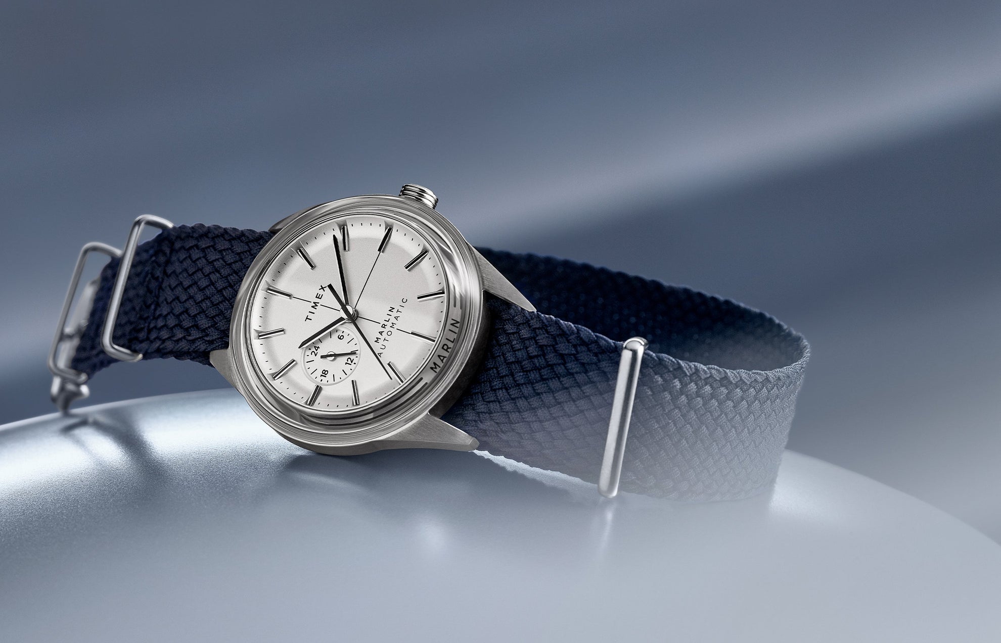 Timex Marlin Jet Automatic watch with silver-tone dial, navy woven strap, and stainless-steel case on soft blue gradient background.