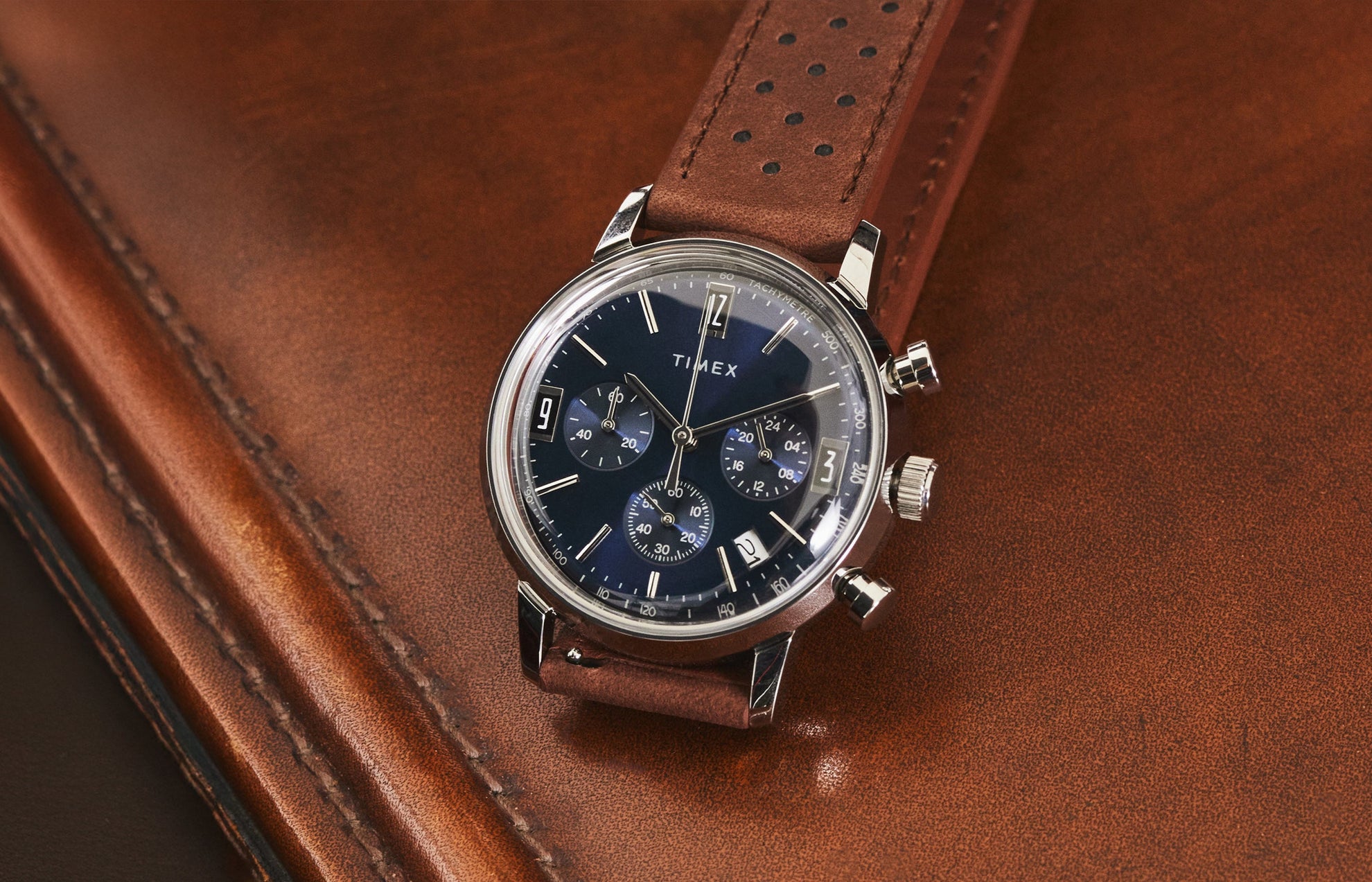 Timex Marlin Chronograph Tachymeter watch with blue dial and brown perforated leather strap resting on brown leather surface.