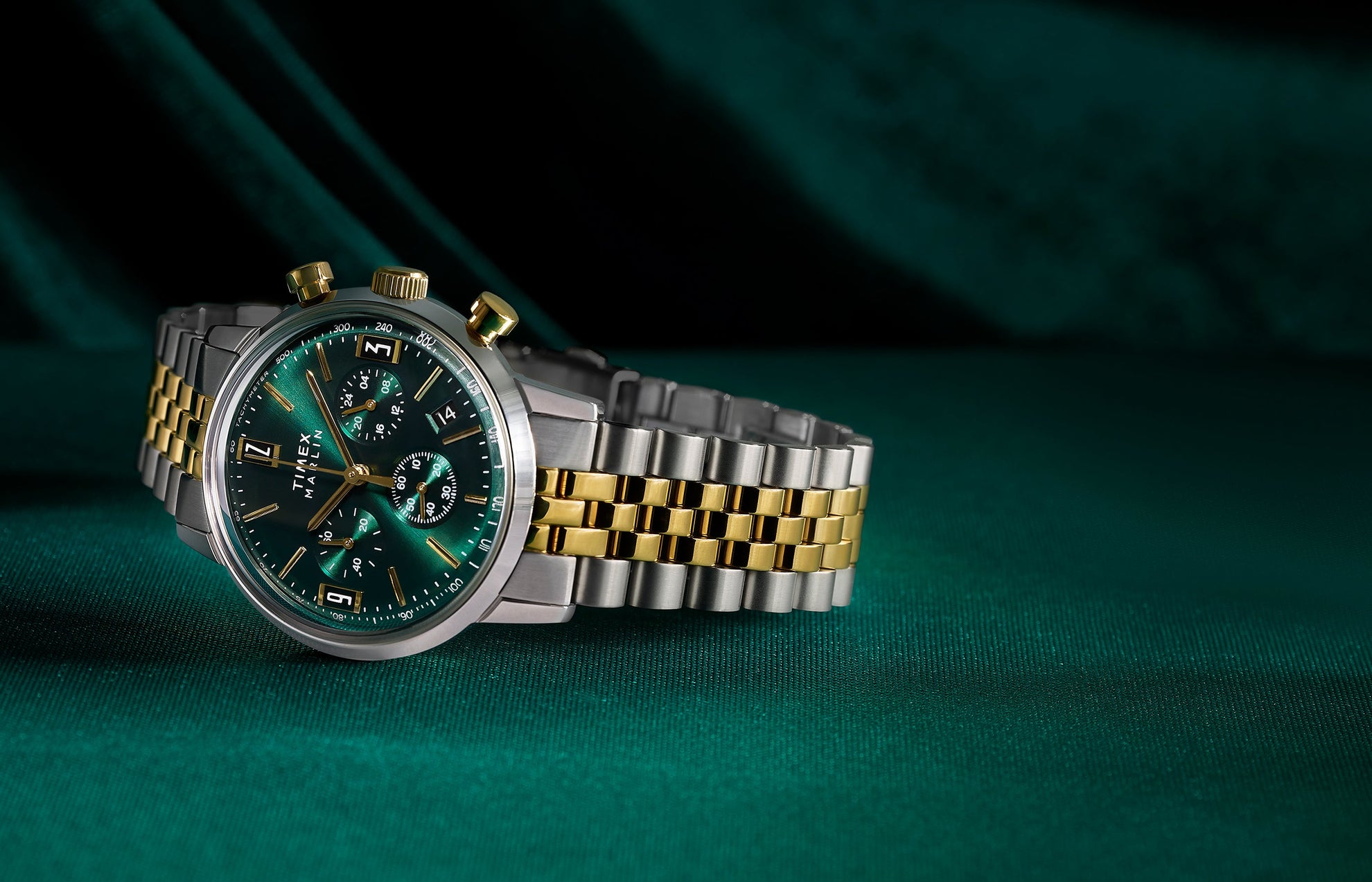 Timex Marlin Chronograph Tachymeter with emerald green dial and gold accents on a two-tone stainless-steel bracelet, displayed on rich green fabric.
