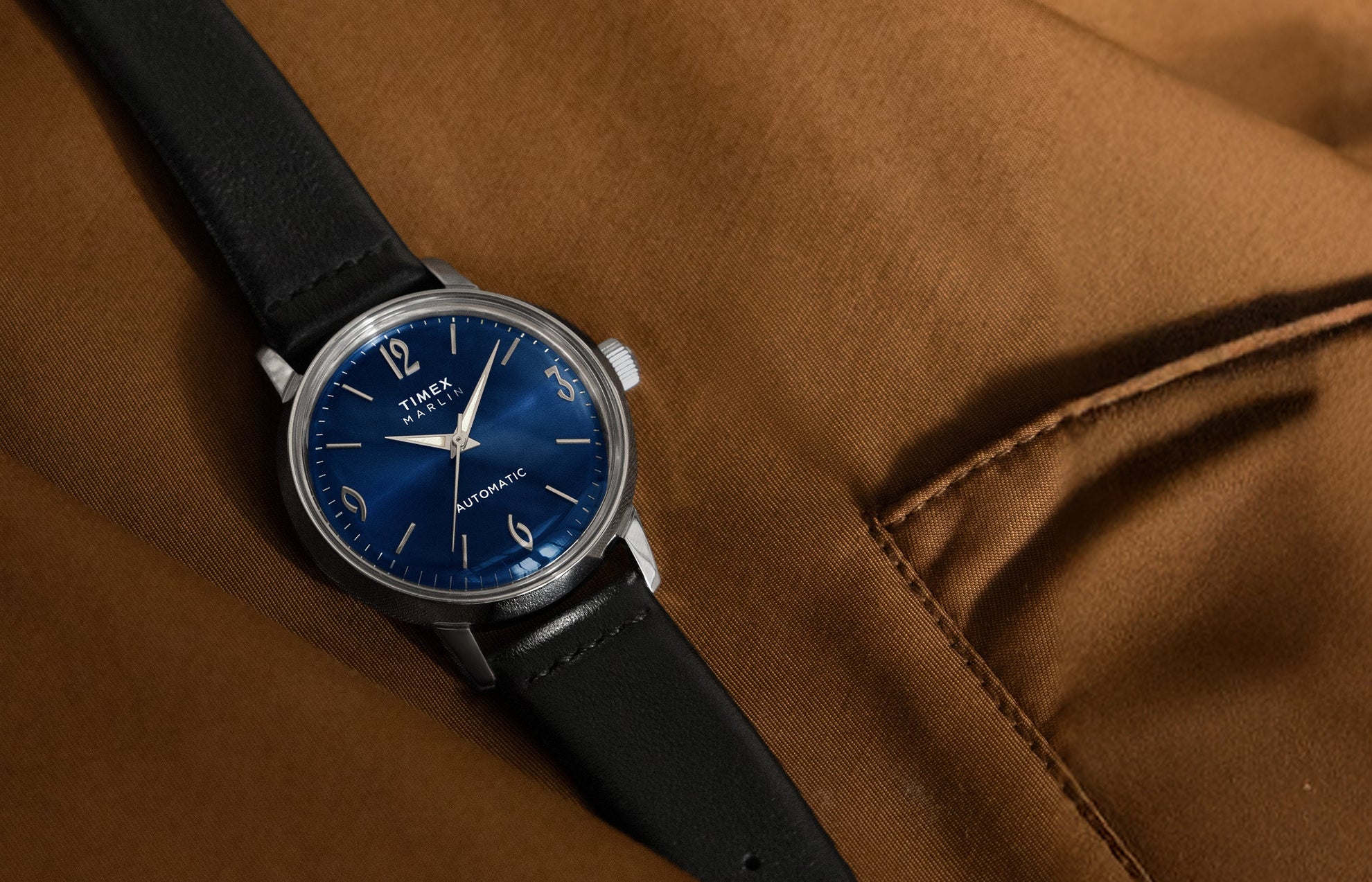 Timex Marlin Automatic watch with deep blue sunray dial and black leather strap resting on a brown fabric surface.