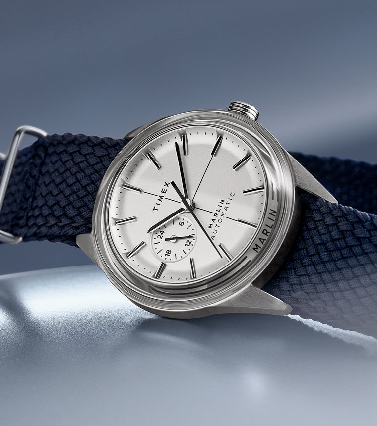 Timex Marlin Jet Automatic watch with silver-tone dial, navy woven strap, and stainless-steel case on soft blue gradient background.