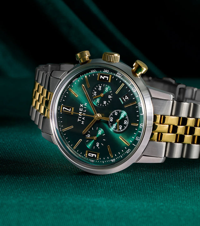 Timex Marlin Chronograph Tachymeter with emerald green dial and gold accents on a two-tone stainless-steel bracelet, displayed on rich green fabric.