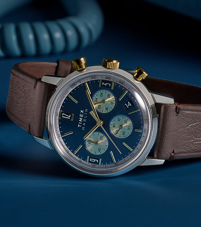 Timex Marlin Chronograph Tachymeter with blue dial and gold accents worn on wrist, styled with burgundy suit jacket and white cuff.