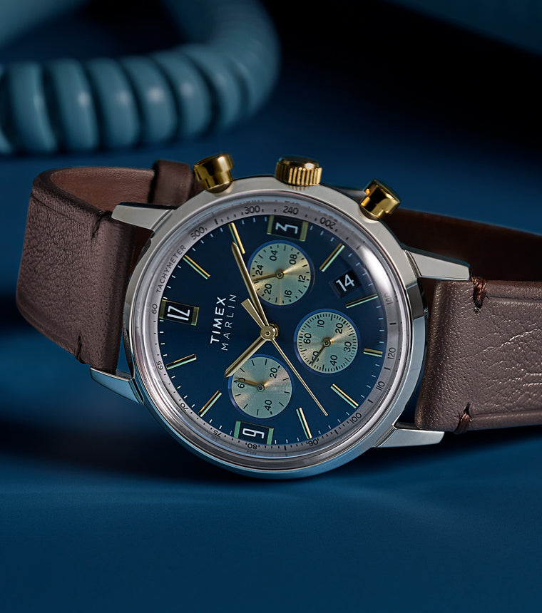Timex Marlin Chronograph Tachymeter with blue dial and gold accents worn on wrist, styled with burgundy suit jacket and white cuff.