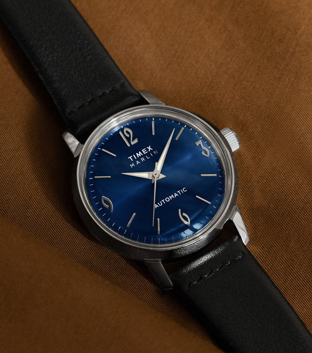 Timex Marlin Automatic watch with deep blue sunray dial and black leather strap resting on a brown fabric surface.